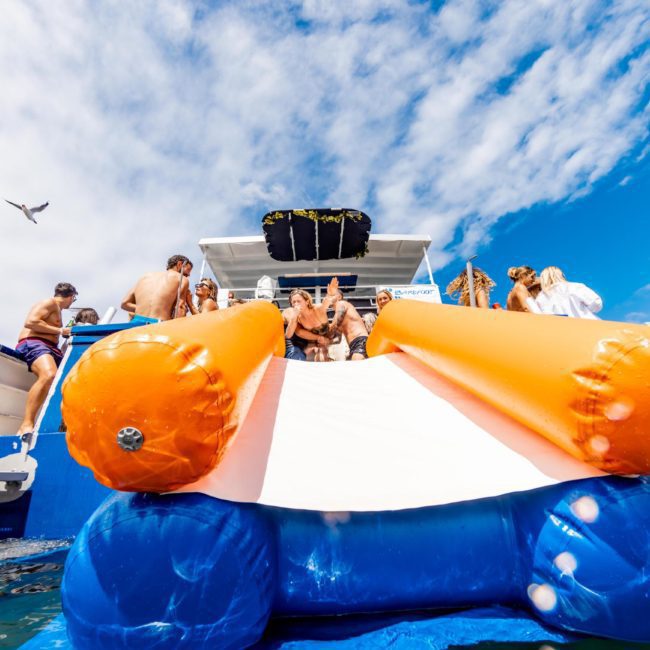 A group of people are on a boat with an inflatable slide extending into the water, against a backdrop of blue sky and clouds with birds flying. Experience ultimate fun with our DJ boat hire Sydney service, perfect for any occasion.