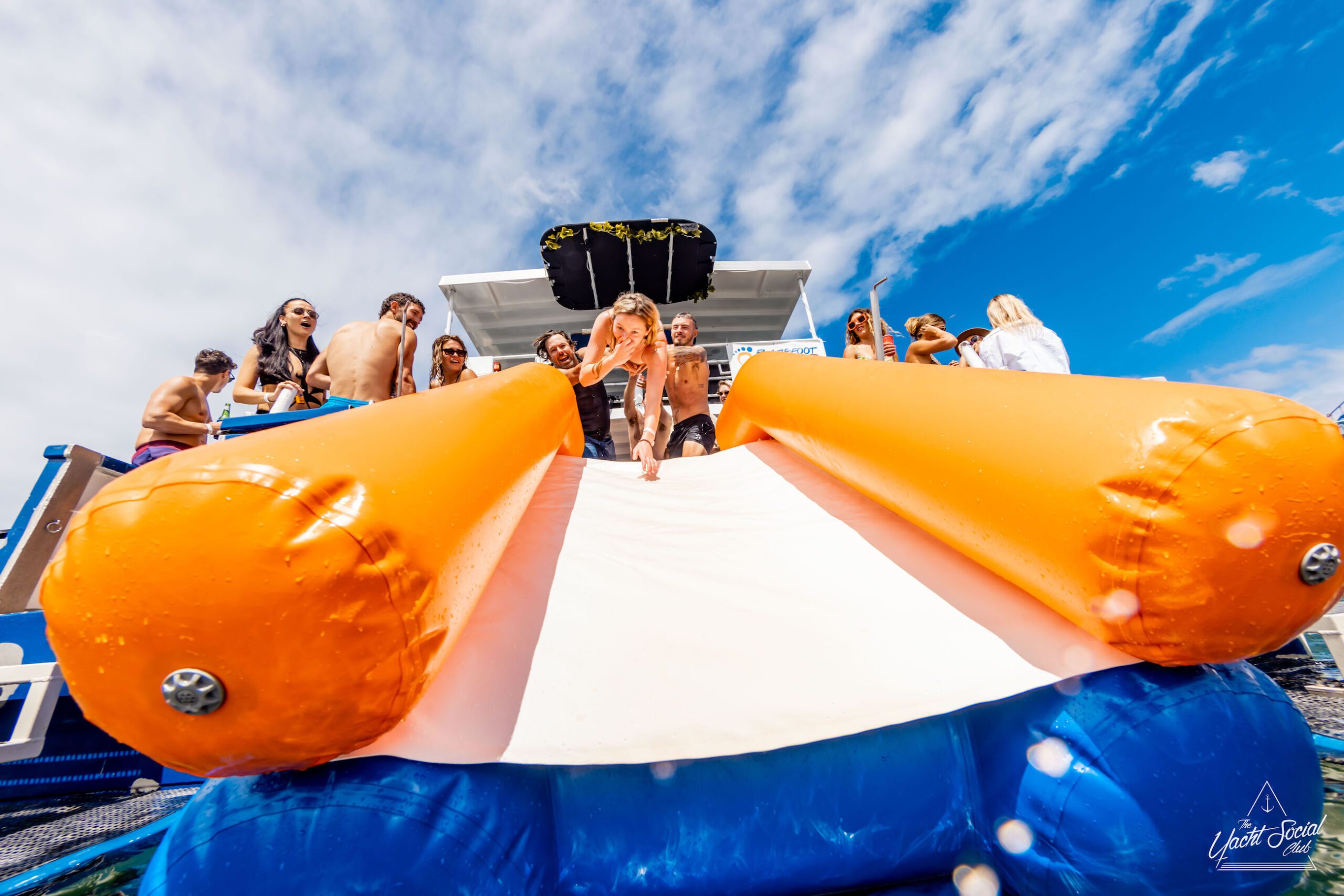 A group of people enjoys a sunny day on a DJ boat hire in Sydney, equipped with a large inflatable slide. One person is sliding down the bright orange and white slide into the water.