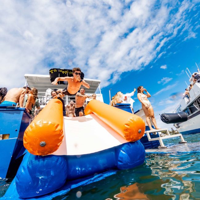 A person slides off an inflatable slide into the ocean from a boat, surrounded by others on a sunny day with clear skies. It’s the perfect scene for a Sydney boat party hire, complete with laughter and fun.