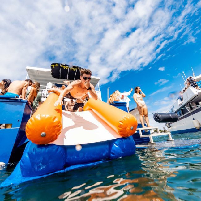 A man is sliding off an inflatable slide from a boat into the water, surrounded by people in swimwear. The scene takes place on a sunny day with other boats and clear skies visible in the background, capturing the essence of a lively Sydney boat party hire.