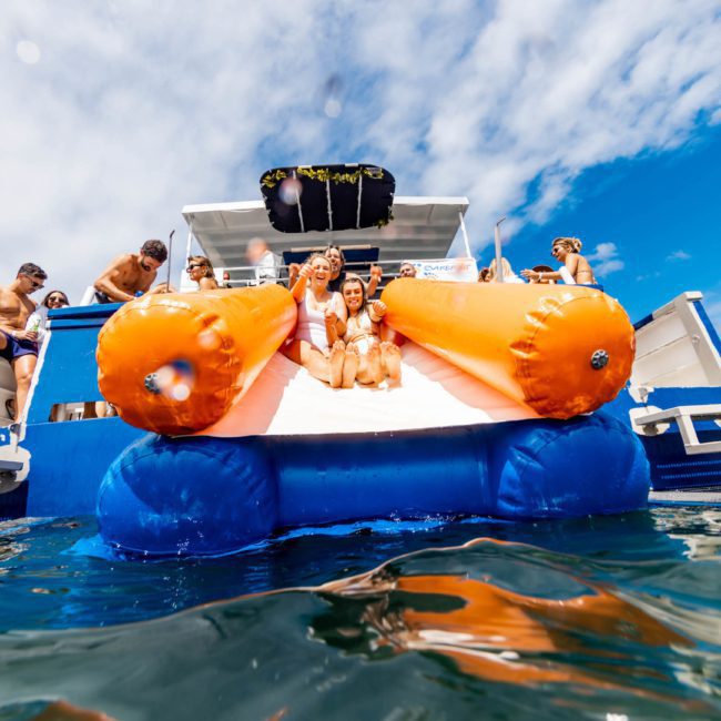 People on a catamaran party in Sydney enjoy a water slide leading into the ocean on a sunny day, with some individuals preparing to enter the water and others watching from the boat.