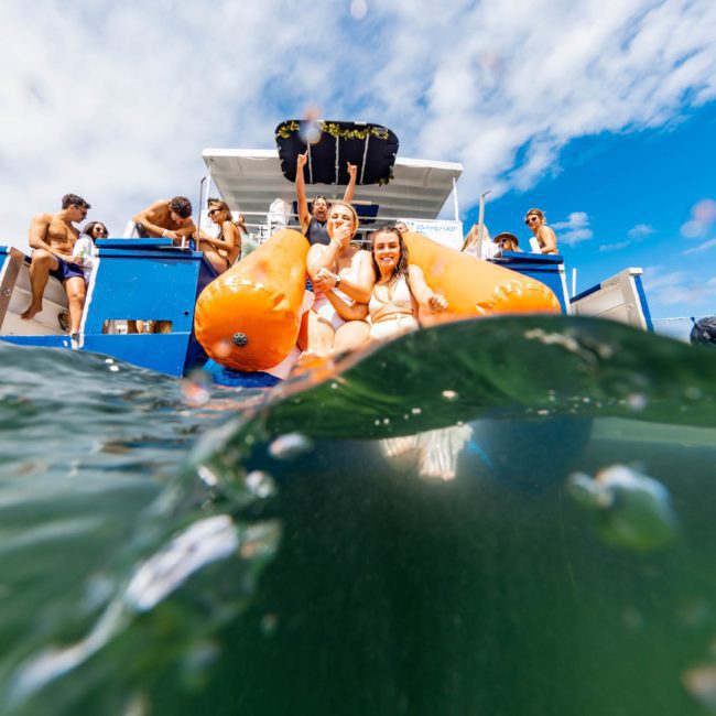People aboard a boat enjoy themselves in the water, with two individuals sitting on an orange inflatable float. The sky is partly cloudy, and others are visible on the boat’s upper deck, experiencing a private yacht charter in Sydney Harbour.
