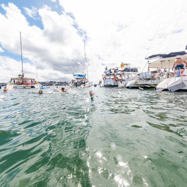 People are swimming and lounging on boats in a sunny, crowded marina with sailboats, motorboats, and luxury yacht hire Sydney options.
