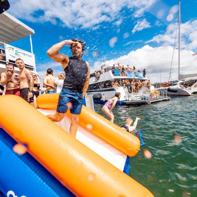 A man strikes a pose on an inflatable slide at a water party with boats and onlookers in the background, enjoying the festive vibes of a catamaran party Sydney.