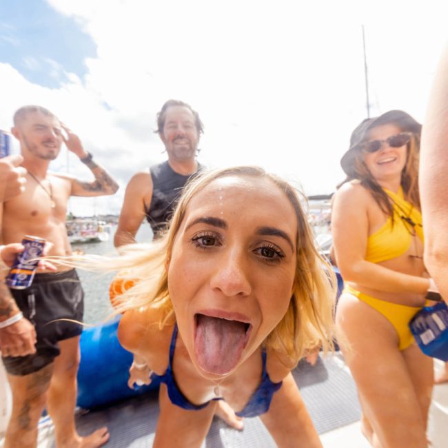 A group of people in swimsuits are enjoying a sunny day on a private yacht charter in Sydney Harbour. One woman is sticking her tongue out at the camera, while others are smiling and holding drinks.