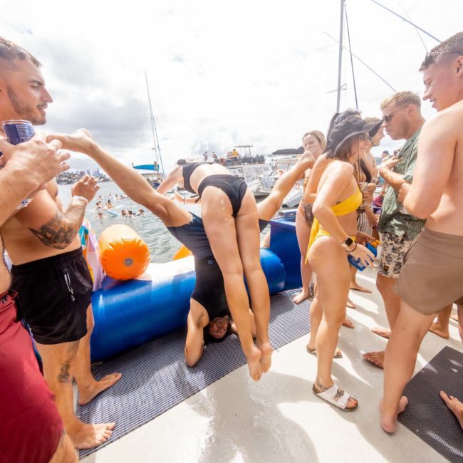 A group of people on a private yacht charter in Sydney Harbour gather around a person performing a headstand on a flotation device, with drinks in hand. Other boats and people are visible in the background on a sunny day.