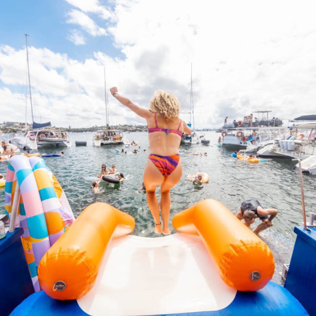 A woman in a bikini jumps off the back of a boat into the water during a lively Sydney boat party. Several boats and people are in the background on a sunny day.