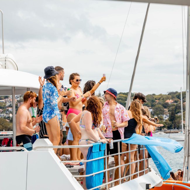 A group of people in swimwear and casual attire are gathered on the deck of a luxury yacht in Sydney, socializing and holding drinks, with a hilly coastline and houses visible in the background.