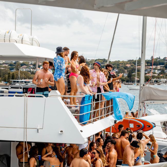 A group of people in swimsuits and casual clothing gather on the upper and lower decks of a boat, possibly at a Catamaran party Sydney with a shoreline and other boats visible in the background.