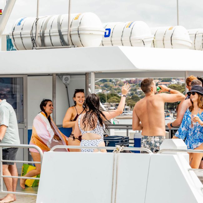 A group of people in swimwear are socializing and dancing on the deck of a catamaran on a sunny day, enjoying what feels like the perfect Sydney boat party hire experience.