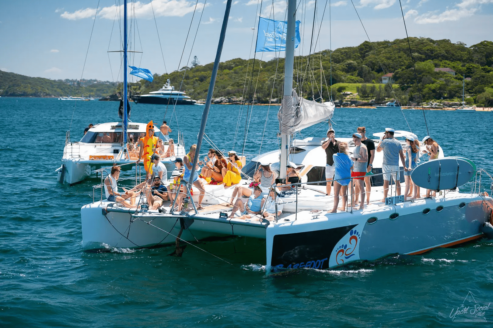 A group enjoys relaxing and socializing on the Barefoot catamaran, anchored in sunny blue water near a forested shoreline, with another boat and yachts in the background.