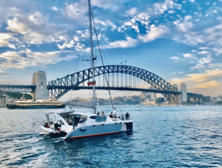 A white sailboat named Barefoot, with people onboard, sails on a blue harbor with the Sydney Harbour Bridge and city skyline in the background beneath a partly cloudy sky.