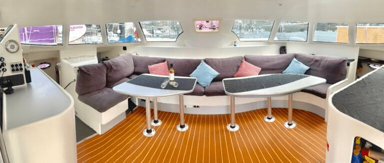 The interior of the Barefoot lounge features curved gray couches, three tables with a bottle and glasses, colorful throw pillows, large windows, and striped wood flooring.