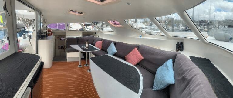 Barefoot features a modern interior with large windows, grey cushioned seating, colorful throw pillows, and two small tables. The bright cabin is filled with natural light and offers stunning marina views.