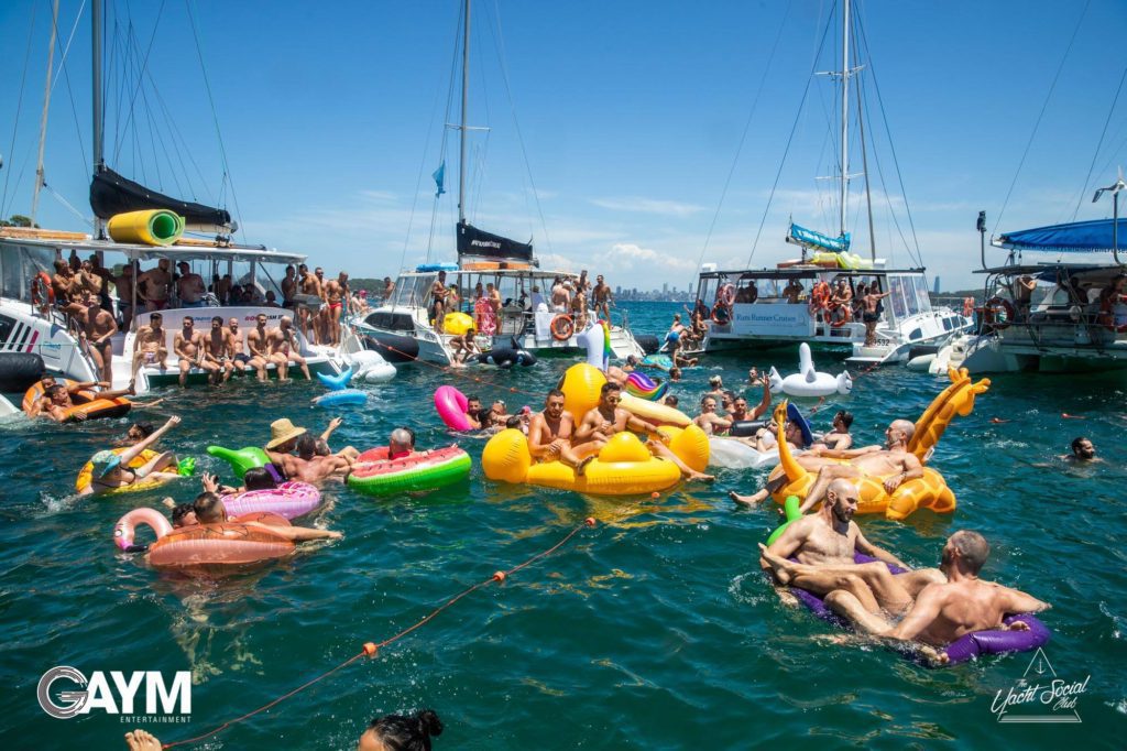 People are enjoying a sunny day in the water with inflatable floats. Boats are anchored nearby, and there are groups of people socializing both on the boats and in the water during what appears to be a luxury yacht hire Sydney event.