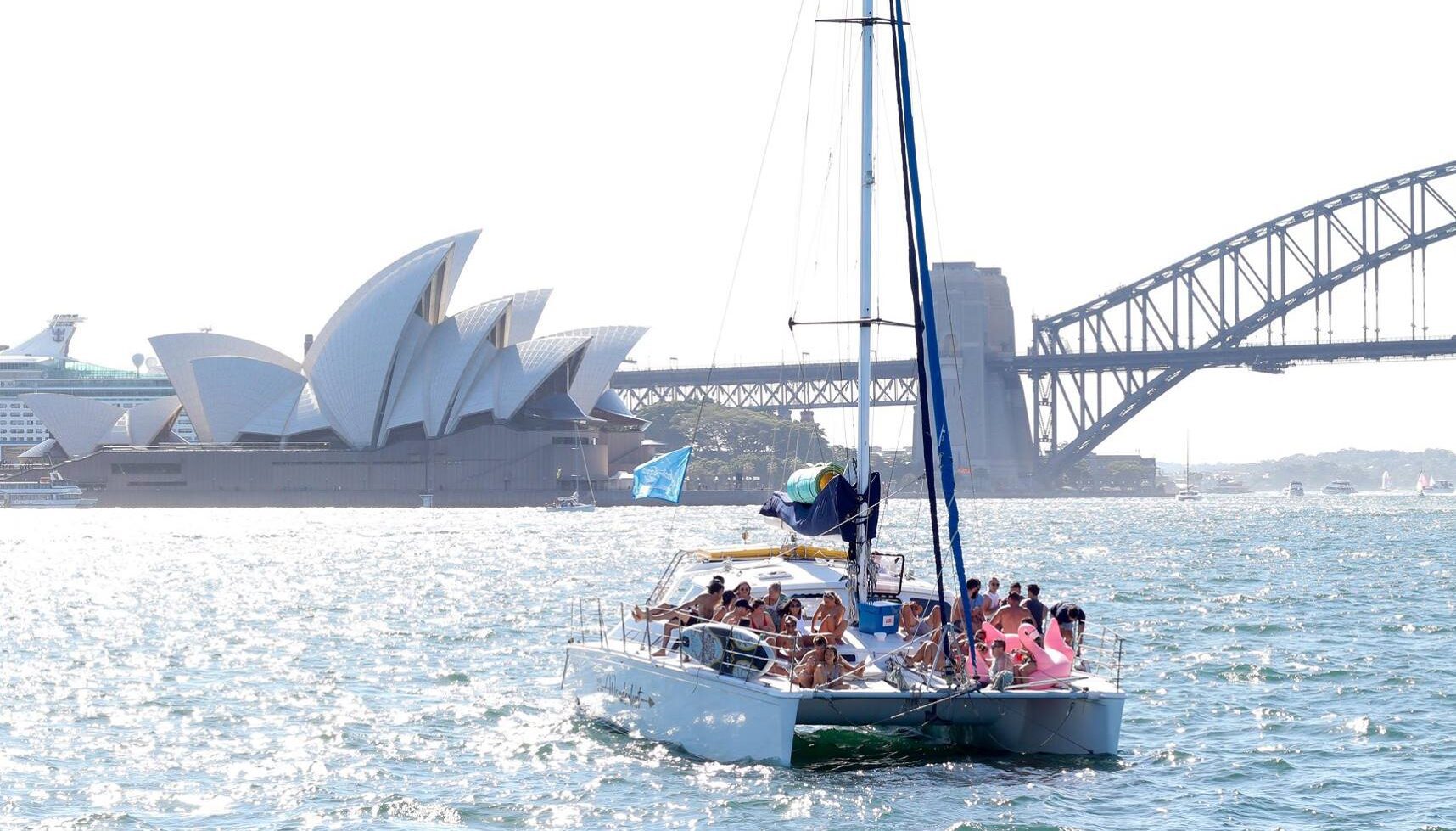 A sailboat filled with people navigates waters near the Sydney Opera House and Sydney Harbour Bridge on a bright day, perfect for a luxury yacht hire Sydney.