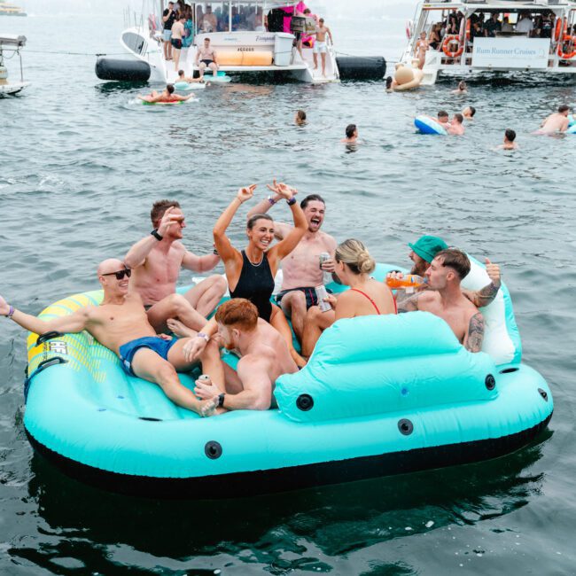 A group of eight people are having a blast on an inflatable raft in the middle of the water. They are smiling, raising their arms, and cheering with joy. Other rafts and boats with more people add to the lively, festive atmosphere in the background.