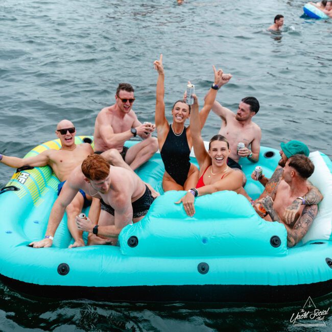 A group of seven people enjoying themselves on a large inflatable raft in a body of water. They are smiling, holding drinks, and wearing swimwear. One woman in the center is raising her arms in excitement. Music can be heard from other rafts and people visible in the background.