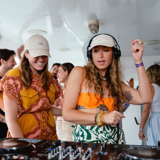 People on a boat party, with a woman in the foreground wearing headphones and a colorful outfit DJing on a mixer. Another woman beside her, wearing a hat and patterned dress, watches attentively. Others in the background are socializing and enjoying the beach-themed atmosphere. Life jackets hang on the wall.