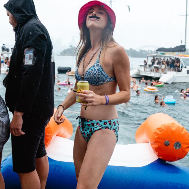 A woman sporting a pink bucket hat and bikini, with a fake mustache on her face, stands on a large inflatable flamingo floating in the water. She holds a drink in one hand. People are swimming and partying on boats in the background under a cloudy sky.