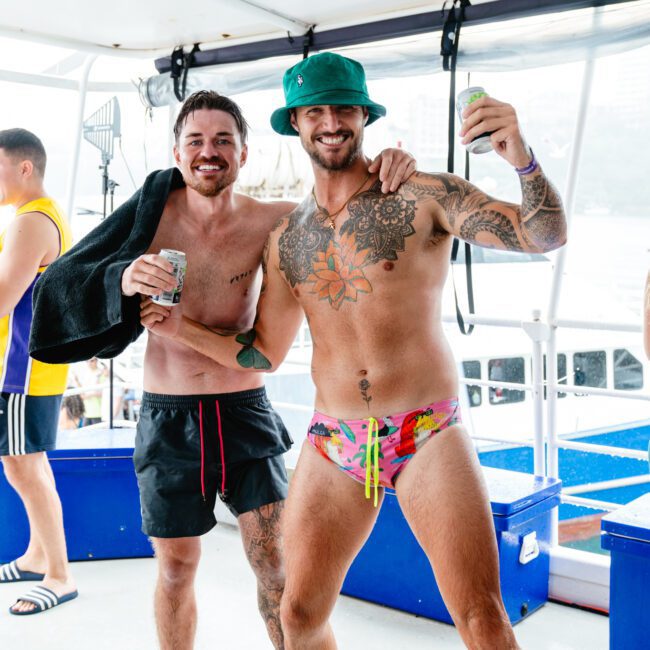 Two men on a boat, smiling and posing with drinks. One in black swim shorts has a towel draped over his shoulder, while the other wears a green bucket hat and floral swim briefs. Both have tattoos and appear to be enjoying themselves on the water.