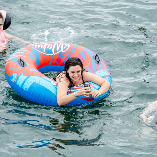Three people are in the water, enjoying a swim. A woman holds onto a colorful inflatable ring while smiling brightly. They are surrounded by two men, one clutching a vibrant beach ball, and another floating nearby, looking cheerful. An additional inflatable ring floats in the background.