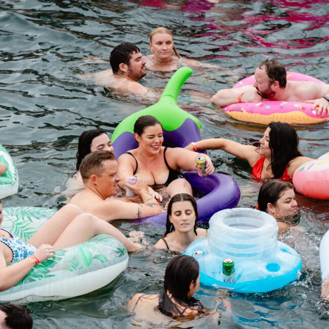 A group of people enjoying a swim in the water, lounging on various inflatable floats, including a donut and an avocado. Some are holding drinks and smiling, creating a festive and relaxed atmosphere under the sun.