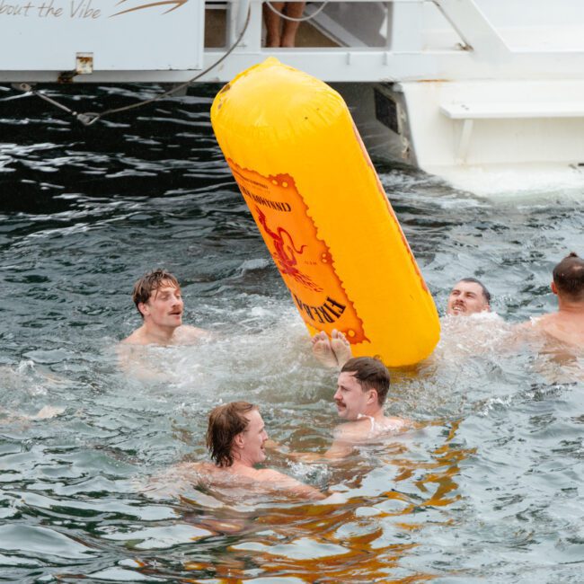 A group of six men in swim trunks are playing in the water near a boat. They are gathered around a large yellow inflatable buoy, laughing and splashing. The boat's platform is visible in the background. The dark water suggests it might be a lake or the ocean, setting a fun summertime scene.


Keywords: summertime