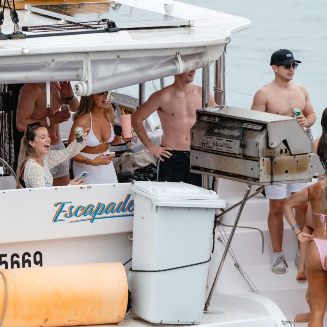 A group of people are gathered on a boat named "Escapade." Some are sitting, while others are standing; one is grilling on a barbecue. Several are in swimwear, enjoying drinks. A yellow float and an inflatable flamingo add color to the water.