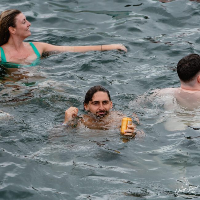 A man with long hair is holding a can while swimming in the water with two other people. The woman on the left, wearing a green swimsuit and smiling, enjoys the slightly choppy water. The person on the right, with short dark hair, is seen from behind.
