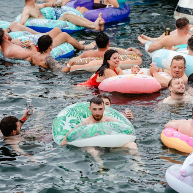 A group of people relaxes on inflatable pool floats with various designs while enjoying drinks in the water. There are several individuals socializing and having fun in the vibrant outdoor aquatic setting, creating a lively atmosphere.