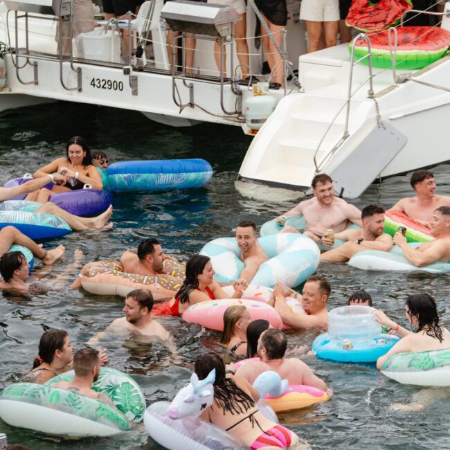 A lively scene of a pool party on water, featuring numerous people in swimsuits enjoying themselves on inflatable floaties. Behind, a crowded yacht has more people mingling and observing the fun, with one inflatable watermelon prominently in the background.