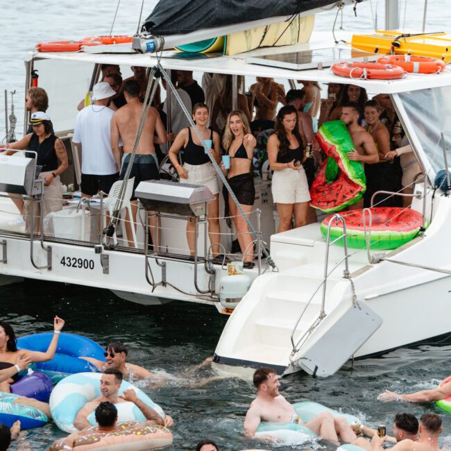 A lively boat party is captured in the image. The boat is crowded with people dancing and mingling, while several individuals in colorful floats enjoy the water nearby. The atmosphere is festive, with a mix of inflatable drinks holders and vibrant floaties adding to the fun blissful scene.