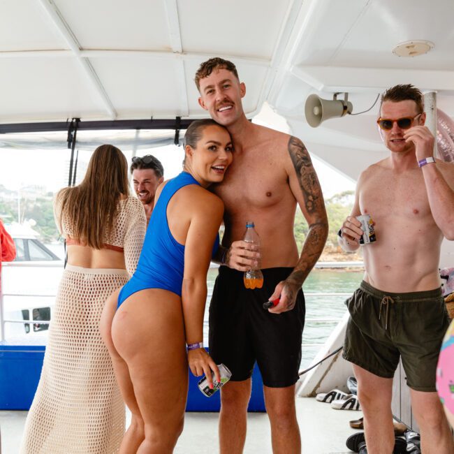 A group of people on a boat enjoying a party. A woman in a blue swimsuit and a man with tattoos, both smiling at the camera while holding drinks, are at the center. Others around them are casually dressed, with some capturing photos. The setting is relaxed and festive.
