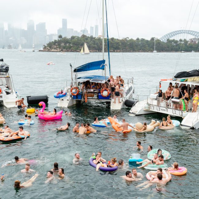 A lively scene on a river with many people swimming and floating on inflatable toys between several boats. The city skyline, including an iconic bridge, is visible in the background under a cloudy sky. The boats have groups of people on board, appearing festive and relaxed.