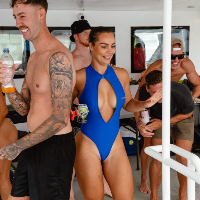 A group of people are enjoying themselves on a boat. A smiling man in black shorts and a woman in a blue one-piece swimsuit are in the foreground, both holding drinks. The water glistens under the bright sun. Other people are visible in the background, also laughing and having a great time.