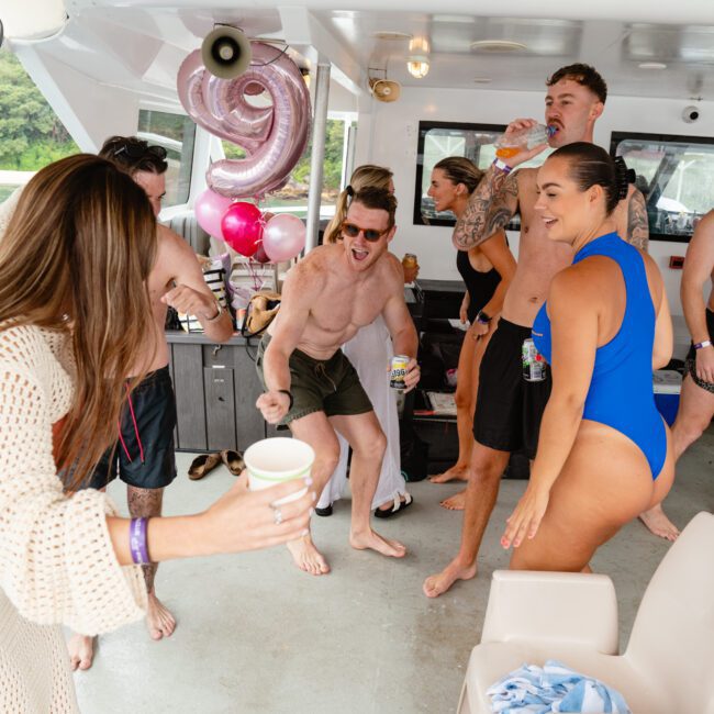 A group of people in swimsuits are partying on a boat, basking in the summer sun. Some are holding drinks, and one person is extending a drink to another. Balloons, including a large "9," float cheerfully in the background. The mood is celebratory and joyful, creating unforgettable memories on the water.