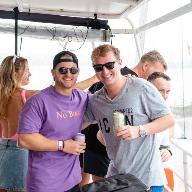 A group of people are on a boat, enjoying what appears to be a social gathering. Two men in the forefront, one wearing a purple "No Buas" t-shirt and sunglasses, the other in a grey "College 22" t-shirt, smile at the camera while others mingle in the background amidst a festive atmosphere.