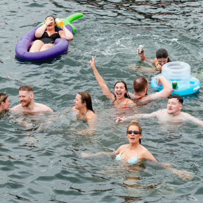 A group of people swimming and relaxing in a body of water. Some are holding drinks and floating on inflatable devices, while others swim or stand in the water. They appear to be enjoying themselves, smiling and making playful gestures amidst the refreshing environment.