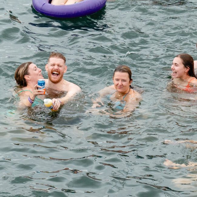 A group of people enjoying a swim in a large body of water. They are smiling, laughing, and holding drinks. One person is lounging on a purple inflatable tube. They all appear relaxed and happy as the water remains clear and slightly wavy, reflecting their joyous spirits in nature's embrace.