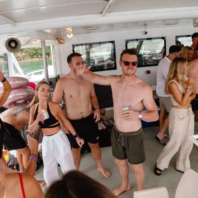 A group of people are enjoying a party on a boat. Some are wearing swimsuits while others are in casual summer attire. The atmosphere is lively, with individuals dancing, holding drinks, and socializing. The boat is docked near a lush, green shoreline under the golden afternoon sun.