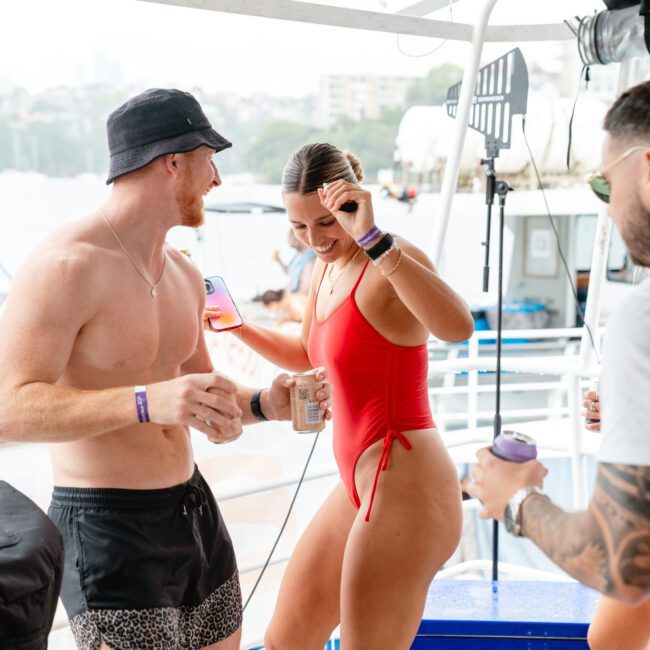 People enjoying a lively outdoor gathering on a boat. A shirtless man in a hat and swim trunks dances with a woman in a red swimsuit, while others stand nearby holding drinks. The background shows an inviting waterfront with buildings and other boats.