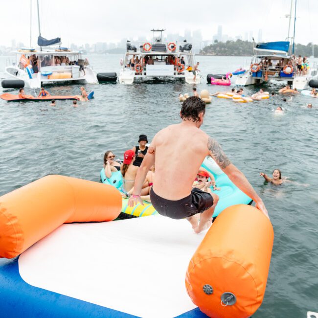 People enjoying a fun day on the water, surrounded by boats and inflatables. A man splashes onto an inflatable mat while others relax and swim around. Overcast skies hint at a possible shower, with a city skyline visible in the background.
