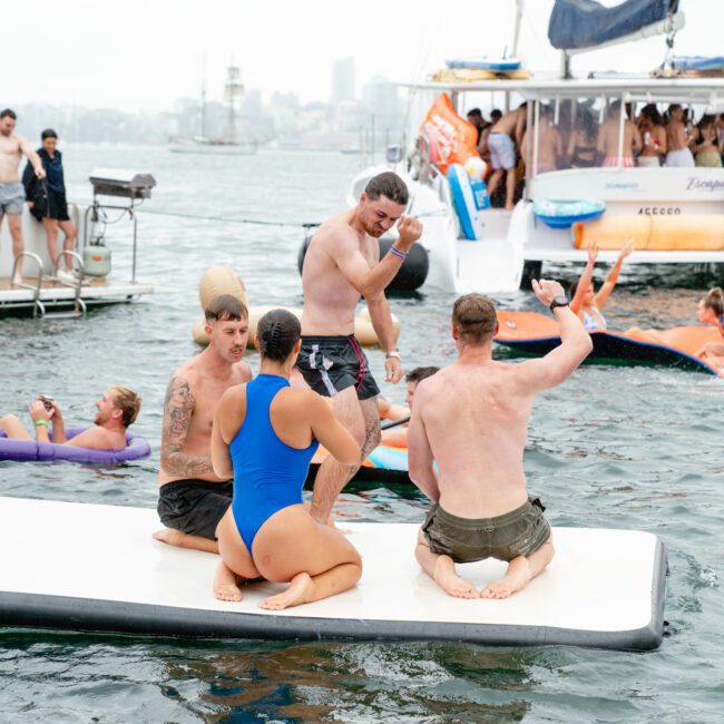 Several people are on an inflated raft in the water, enjoying various activities. One man is standing and talking, two men are seated, and a woman in a blue swimsuit is kneeling. Boats and other people are seen sailing in the background on the water.