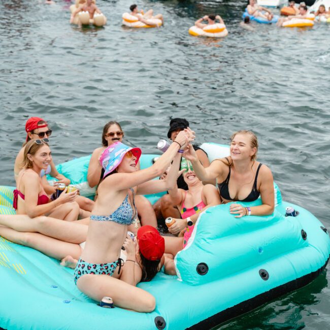 A group of friends enjoying a day on the water. Eight people, wearing swimsuits, are sitting on an inflatable raft and toasting with drinks. Behind them, other people are on smaller floats, and boats are visible in the background, suggesting a vibrant social scene filled with laughter and fun.