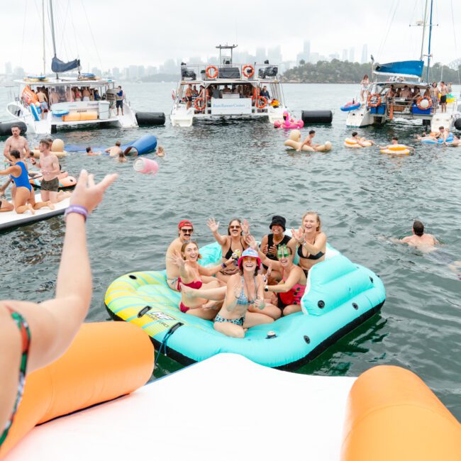 A group of people is having a fun time on an inflatable raft in the water, surrounded by other partygoers on boats and floaties. One person with braided hair is in the foreground, facing the lively gathering. Cityscape and a bridge are visible in the foggy background.