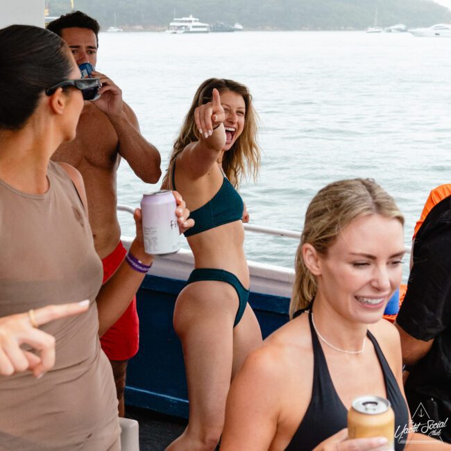 A group of friends enjoying a lively boat party. The woman in the center wearing a teal bikini points towards the camera, smiling. Others around her, dressed in swimwear, hold drinks. In the background, a calm sea with yachts and boats anchored nearby gleams under a hazy sky.