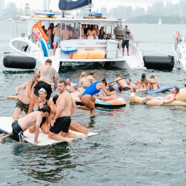 A lively scene of people in swimsuits enjoying a party in the water near a large boat. Some are on colorful floaties, others on paddle boards, and some swimming. There is a cloudy sky and city skyline in the background. The boat is filled with cheerful folks and decorated with vibrant banners.