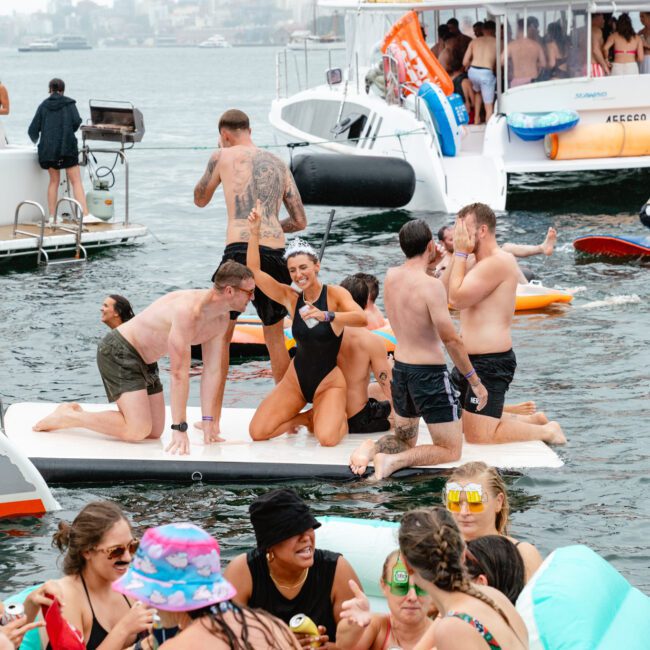 A lively group of people relax and have fun on a floating platform and in the water beside several yachts. Some are dancing, drinking, or chatting, while others float with pool noodles and inflatable toys. The scene depicts a festive atmosphere on a bright afternoon with flowing laughter and music.