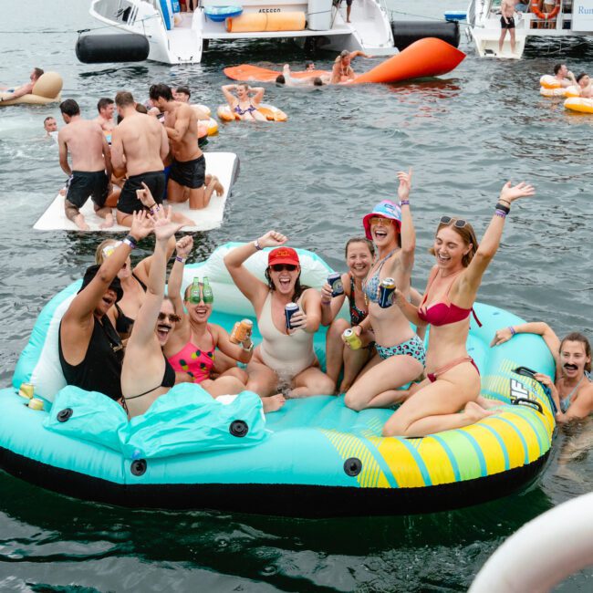 A group of smiling people in swimwear sits on an inflatable raft in a body of water, raising their arms in celebration. Several others are swimming and lounging on floats nearby. Kayaks and more people are in the background, creating a lively party atmosphere.